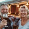 Happy mature couple sitting by their caravan home drinking red wine. Senior husband and wife traveling on holidays by trailer camper van. Monthly Reservation